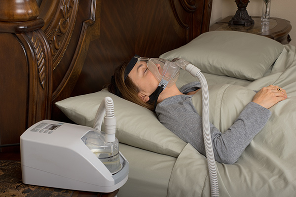 Sleep Apnea in Women vs. Men | Woman sleeping in bed with a mask attached to a CPAP device.